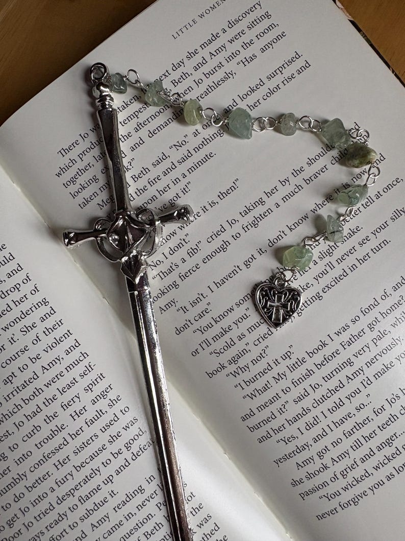 Puede incluir: Un marcap&aacute;ginas plateado con dise&ntilde;o de espada y una cadena de cuentas con piedras verdes descansa sobre un libro abierto. Un dije en forma de coraz&oacute;n con una cruz cuelga de la cadena. El libro est&aacute; abierto en una p&aacute;gina de texto.