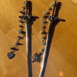 Puede incluir: Dos horquillas plateadas con forma de espada con cadenas decorativas. Una tiene un dije de coraz&oacute;n y cuentas azules, la otra tiene un dije de estrella y cuentas verdes. Los accesorios para el cabello se exhiben sobre una superficie de madera.