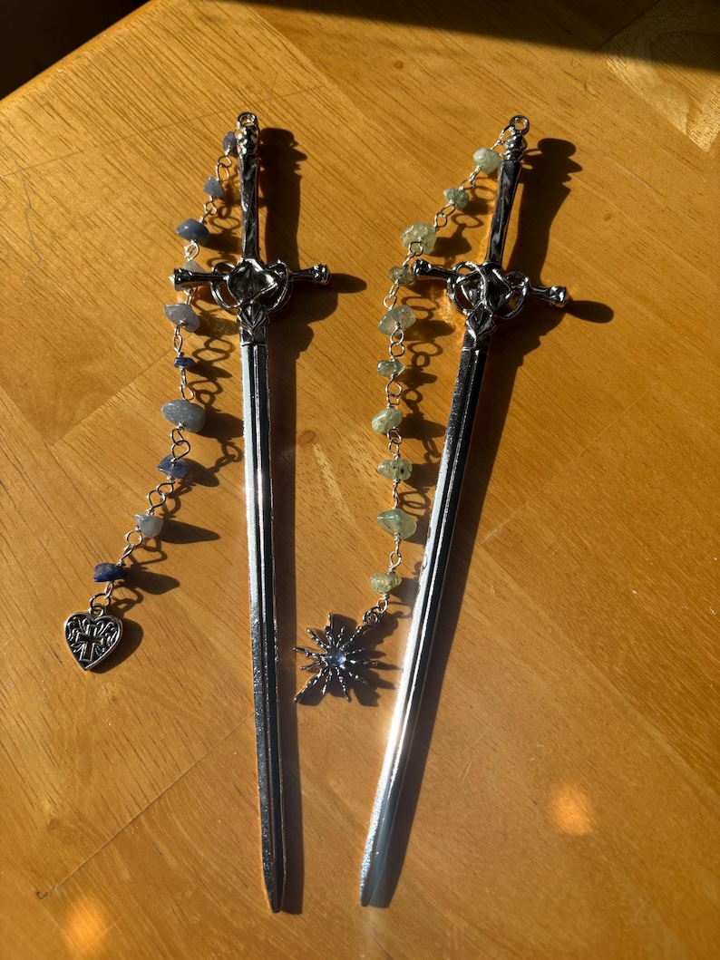 Puede incluir: Dos horquillas plateadas con forma de espada con cadenas decorativas. Una tiene un dije de coraz&oacute;n y cuentas azules, la otra tiene un dije de estrella y cuentas verdes. Los accesorios para el cabello se exhiben sobre una superficie de madera.