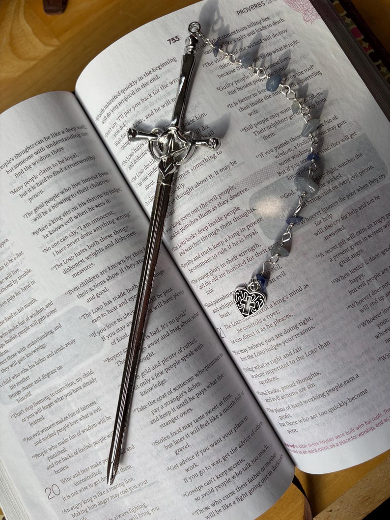 Puede incluir: Un marcap&aacute;ginas plateado con forma de espada y empu&ntilde;adura decorativa descansa sobre un libro abierto. Una cadena de cuentas con piedras azules y un dije de coraz&oacute;n tambi&eacute;n est&aacute; en la p&aacute;gina.
