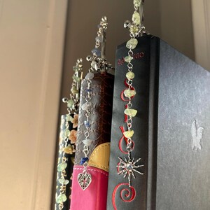 Puede incluir: Primer plano de tres marcap&aacute;ginas decorativos con cuentas de piedras preciosas y dijes de plata. Los marcap&aacute;ginas est&aacute;n insertados en las p&aacute;ginas de libros con cubiertas oscuras. Un marcap&aacute;ginas presenta un dije de coraz&oacute;n con una cruz, otro un sol.