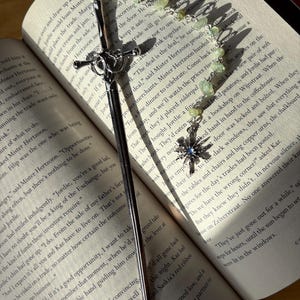 Op de afbeelding: Een zilveren bladwijzer in de vorm van een zwaard met een decoratieve handgreep en een ketting van groene kralen. De bladwijzer ligt op de open pagina's van een boek.