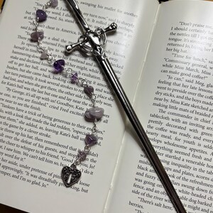 Puede incluir: Un marcap&aacute;ginas plateado con forma de espada, con una empu&ntilde;adura en forma de cruz y una cadena de cuentas de amatista moradas. El marcap&aacute;ginas est&aacute; en un libro abierto.