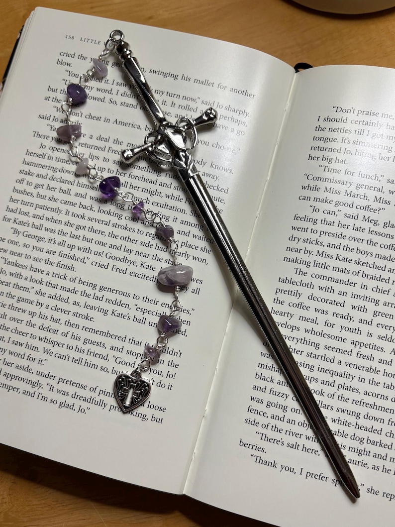 Puede incluir: Un marcap&aacute;ginas plateado con forma de espada, con una empu&ntilde;adura en forma de cruz y una cadena de cuentas de amatista moradas. El marcap&aacute;ginas est&aacute; en un libro abierto.