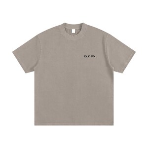 May include: A short-sleeved, taupe-colored t-shirt with the text "SOLID TEN" printed in black on the upper chest. The shirt has a crew neck and a relaxed fit. The fabric appears to be a soft, cotton blend.