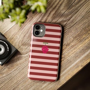 Peut inclure: Une coque de smartphone avec un motif de rayures horizontales alternant des nuances de rouge foncé et de rose clair. Au centre du dos de la coque se trouve un petit graphique stylisé d'une framboise avec une tige et une feuille vertes.