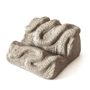 Op de afbeelding: Een grijze betonnen sculptuur van een opgerolde slang. Het lichaam van de slang is getextureerd met schubben en krult rond een rechthoekige basis. De sculptuur heeft een neutrale grijze kleur en staat op een witte achtergrond.