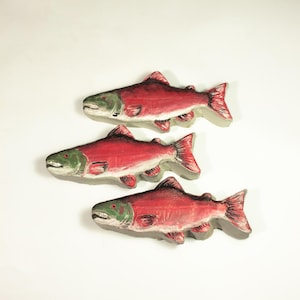 May include: Three decorative fish sculptures. Each fish is painted with a red body, gray underbelly, and green head. The fish are arranged in a vertical stack against a white background.