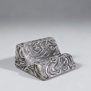 Può includere: Un supporto per telefono color argento con un motivo a spirale unico in bianco e nero. Il supporto è rettangolare con una fessura per tenere un telefono e un supporto posteriore. Il design ricorda le venature del legno.