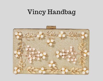 May include: Gold rectangular clutch with floral and pearl embellishments. The clutch features a glittery surface and is adorned with gold-colored leaves and white pearls. The text "Vincy Handbag" is displayed at the top.