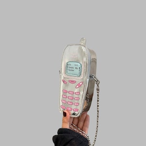 Pode incluir: Uma bolsa transversal prateada metalizada em formato de telemóvel vintage. A bolsa apresenta um ecrã azul claro com o texto Delete My Number e botões cor-de-rosa. Inclui uma alça de corrente prateada e um fecho de correr lateral.