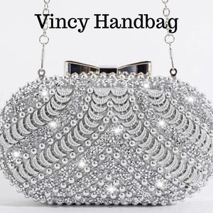 Op de afbeelding: Zilveren clutch handtas met een striksluiting en kettingriem. De handtas is bedekt met kleine, sprankelende versieringen en de tekst "Vincy Handbag" staat bovenaan.