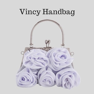 May include: A Vincy Handbag with a silver handle and clasp. The handbag is decorated with several light purple fabric roses. The bag has a silver frame and a light gray body. The text "Vincy Handbag" is above the bag.