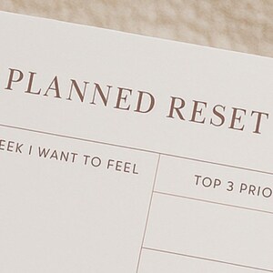 Könnte beinhalten: Nahaufnahme einer Planerseite mit den Worten "PLANNED RESET" in brauner Schrift. Darunter steht der Text "WEEK I WANT TO FEEL" und "TOP 3 PRIORITIES". Die Seite ist hellbeige.