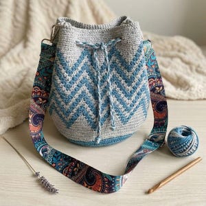 May include: A grey and blue crochet bucket bag with a chevron design. The bag features a patterned strap and a drawstring closure. A ball of yarn and a crochet hook are also visible.