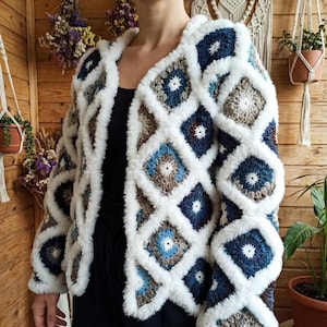 Puede incluir: Un cárdigan hecho a mano, sin cierre, con un diseño geométrico único. La chaqueta presenta un patrón de cuadrados azules, marrones y blancos, delineados con un ribete blanco esponjoso. Esta prenda acogedora y texturizada es una adición elegante a cualquier guardarropa.
