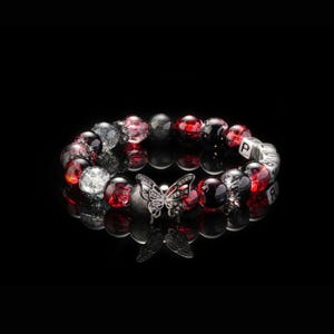 May include: A beaded bracelet featuring a butterfly charm. The bracelet has red, black, and clear beads. The butterfly charm is silver with clear accents. The bracelet is displayed on a reflective black surface.