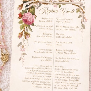 Può includere: Un rosario con perline rosa e un crocifisso e medaglione color oro. Una carta con il testo "Regina Caeli" e illustrazioni floreali è accanto al rosario. La carta ha anche testo in latino e inglese.