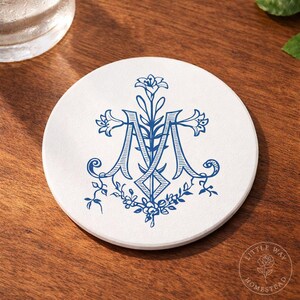 Puede incluir: Un posavasos redondo blanco con un diseño de monograma floral azul. El monograma, la letra "M", está adornado con elementos florales y un borde floral. El posavasos está sobre una superficie de madera.