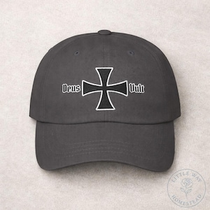 May include: A dark grey baseball cap with a white and black cross emblem and the words "Deus Vult" embroidered above and to the right of the cross. The cap has a curved peak and a button on top.