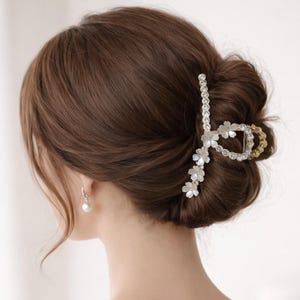 May include: A silver and gold hair claw clip with a floral design, holding up brown hair in a bun. The clip features clear crystals and small white flowers. A pearl earring is visible.