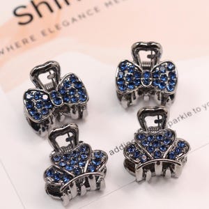 May include: Four small hair claw clips made of dark metallic material, each featuring a bow-shaped design encrusted with numerous small, bright blue rhinestones. The clips are arranged on a white card with the text ShineEve visible in the corner.