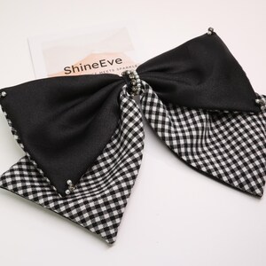 Puede incluir: Un lazo a cuadros blanco y negro con un centro negro, adornado con pequeñas cuentas plateadas. El lazo está en capas, con el texto "ShineEve" visible en una tarjeta blanca en el fondo. Un elegante accesorio para el cabello.