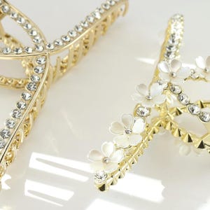 May include: Two gold-colored hair claw clips. One clip is adorned with clear rhinestones, while the other features white flower accents and rhinestones. These decorative hair accessories are designed to hold hair in place.