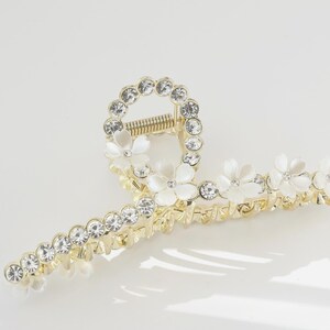 May include: Gold-toned hair claw clip adorned with clear rhinestones and white flower accents. The clip features a circular design with rhinestones and a row of rhinestones along the clip's arms. The claw clip is designed to hold hair in place.