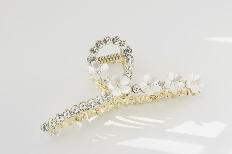 May include: Gold-toned hair claw clip adorned with clear rhinestones and white flower accents. The clip features a circular design with rhinestones and a row of rhinestones along the clip's arms. The claw clip is designed to hold hair in place.
