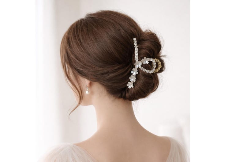 May include: A silver and gold hair claw clip with a floral design, holding up brown hair in a bun. The clip features clear crystals and small white flowers. A pearl earring is visible.