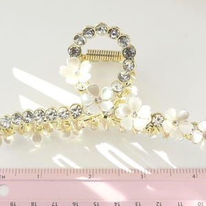 May include: Gold-toned hair claw clip adorned with clear rhinestones and white flower accents. The clip features a circular design with a spring mechanism and measures approximately 4 inches in length.