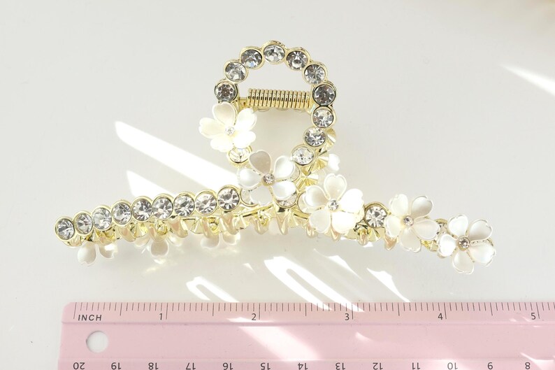 May include: Gold-toned hair claw clip adorned with clear rhinestones and white flower accents. The clip features a circular design with a spring mechanism and measures approximately 4 inches in length.
