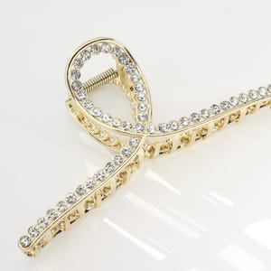 May include: Gold-toned hair claw clip adorned with clear rhinestones. The clip features a decorative design with a spring mechanism. The rhinestones are set along the edges of the clip, adding sparkle and elegance. A stylish hair accessory.