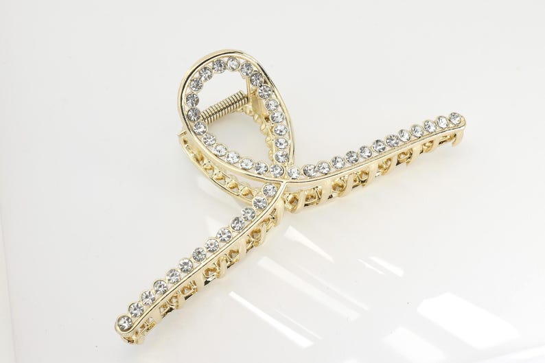 May include: Gold-toned hair claw clip adorned with clear rhinestones. The clip features a decorative design with a spring mechanism. The rhinestones are set along the edges of the clip, adding sparkle and elegance. A stylish hair accessory.