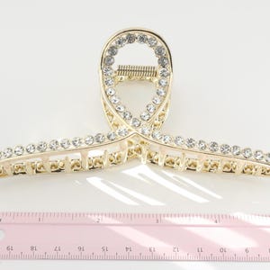 May include: Gold-toned metal hair claw clip with clear rhinestones. The clip is approximately 4 inches long and features a decorative design with a row of rhinestones along the top and bottom edges.