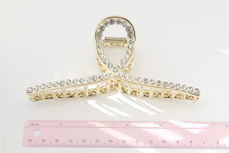 May include: Gold-toned metal hair claw clip with clear rhinestones. The clip is approximately 4 inches long and features a decorative design with a row of rhinestones along the top and bottom edges.