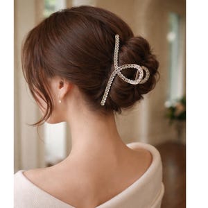 May include: A silver-toned hair claw clip with a decorative, openwork design. The clip is adorned with small, sparkling crystals and is used to secure a bun hairstyle. The hair accessory is a stylish addition to any updo.