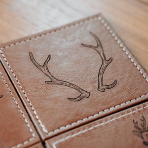 May include: Close-up of a set of brown leather coasters with white stitching. One coaster features a detailed illustration of deer antlers. The coasters are square and appear to be part of a larger set.