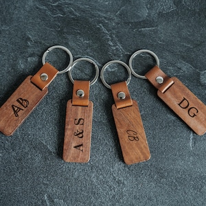 Peut inclure: Quatre porte-clés personnalisés en bois avec des initiales gravées. Chaque porte-clés a une étiquette rectangulaire en bois avec une lanière en cuir et un anneau en métal argenté. Les initiales sont "AB", "A&S", "CB" et "DG".