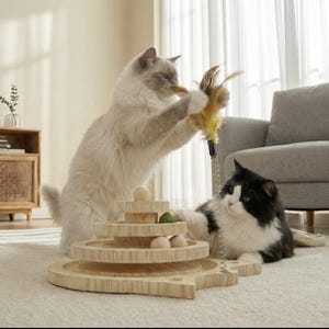 May include: Two cats interacting with a wooden cat toy. One cat is light grey, the other is black and white. The toy features multiple levels with wooden balls and a green ball. A feather toy is being held up.