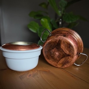 May include: A white ceramic bowl with a copper-colored rim and a copper pot with two handles. The pot is made of copper and has a dark, aged appearance. The bowl is sitting next to the pot on a wooden surface.