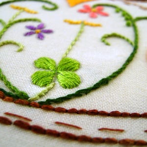 May include: Close-up of an embroidered design featuring a green shamrock, purple flower, and pink flowers on a white fabric background. The embroidery includes green vines and a brown border, showcasing a handcrafted aesthetic.