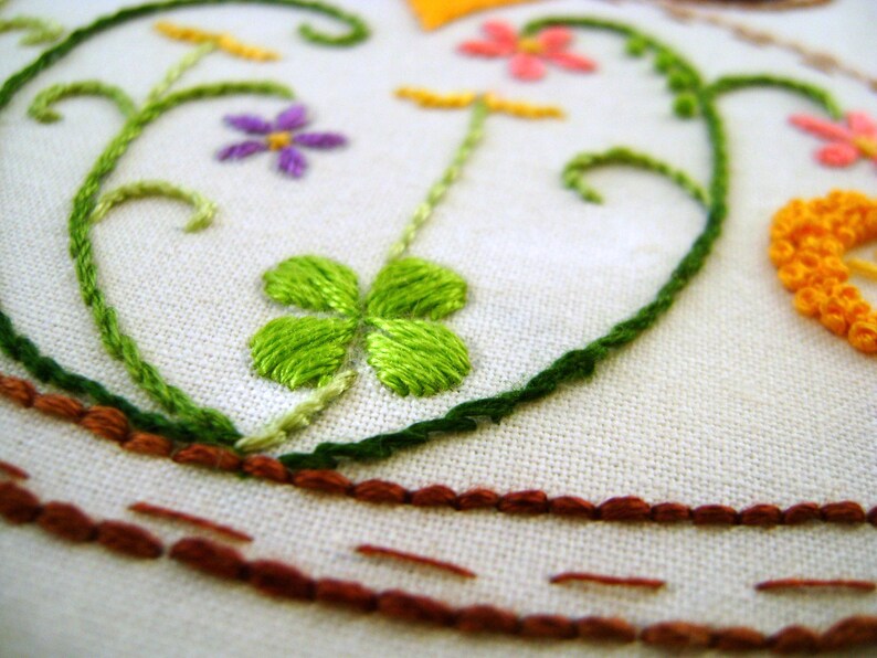 May include: Close-up of an embroidered design featuring a green shamrock, purple flower, and pink flowers on a white fabric background. The embroidery includes green vines and a brown border, showcasing a handcrafted aesthetic.