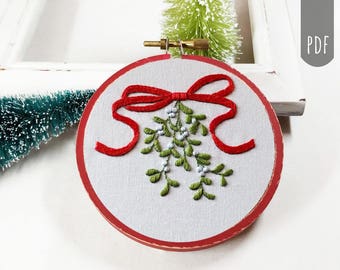 May include: A red and green embroidered hoop art piece featuring a sprig of mistletoe with a red bow. The mistletoe is tied with a red ribbon and has white berries.