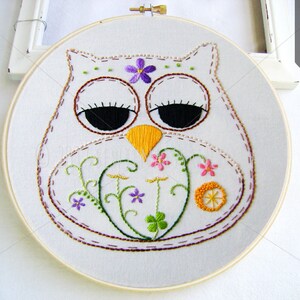 May include: Hand-stitched owl embroidery art on white fabric, set in a wooden hoop. The owl features a white body, brown outline, black eyes, and a yellow beak. Embellished with purple and pink floral details and green vines.