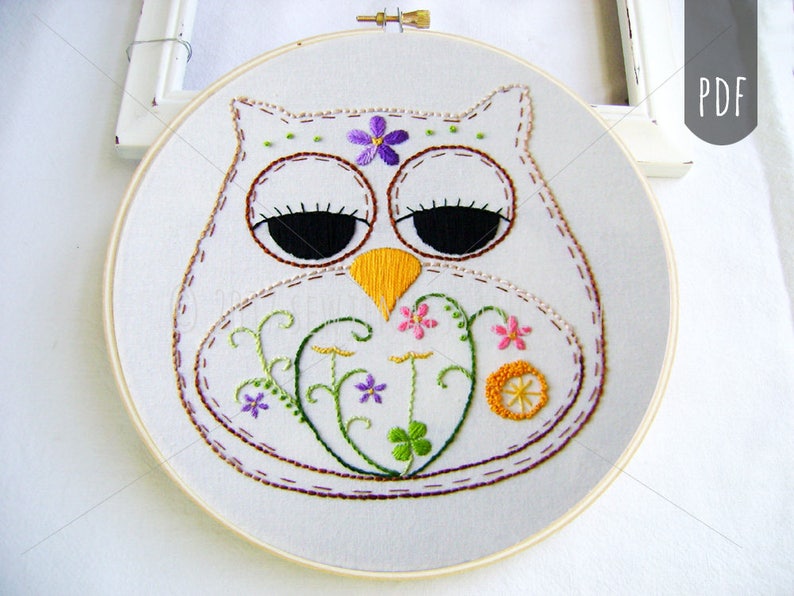 May include: Hand-stitched owl embroidery art on white fabric, set in a wooden hoop. The owl features a white body, brown outline, black eyes, and a yellow beak. Embellished with purple and pink floral details and green vines.