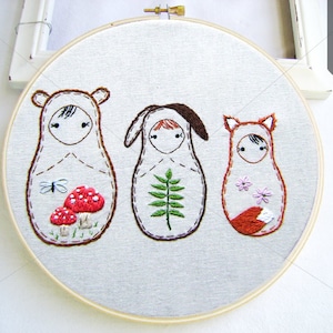 May include: Embroidered hoop art featuring three stylized figures with animal features. The figures are a bear, a deer, and a fox, each with a unique design. The bear has mushrooms, the deer has a fern, and the fox has flowers.