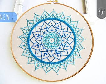 May include: A blue and turquoise mandala embroidery pattern on a white fabric background in a wooden embroidery hoop.