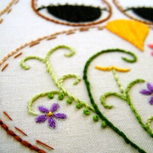 May include: Close-up of a white fabric with embroidered details. The design features an owl with black eyes, a yellow beak, and floral accents in green, purple, and pink. The stitching is in various colors, including brown and orange.